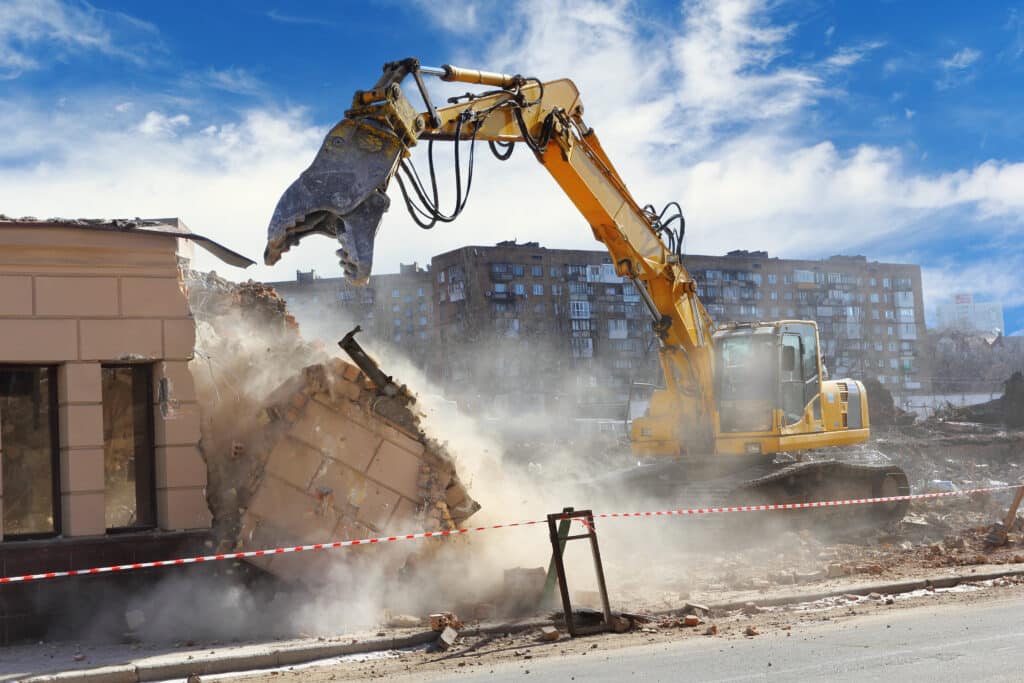 Une excavatrice jaune avec cisailles démolit un bâtiment. Poussière et décombres abondants sur le chantier, ruban de sécurité visible.