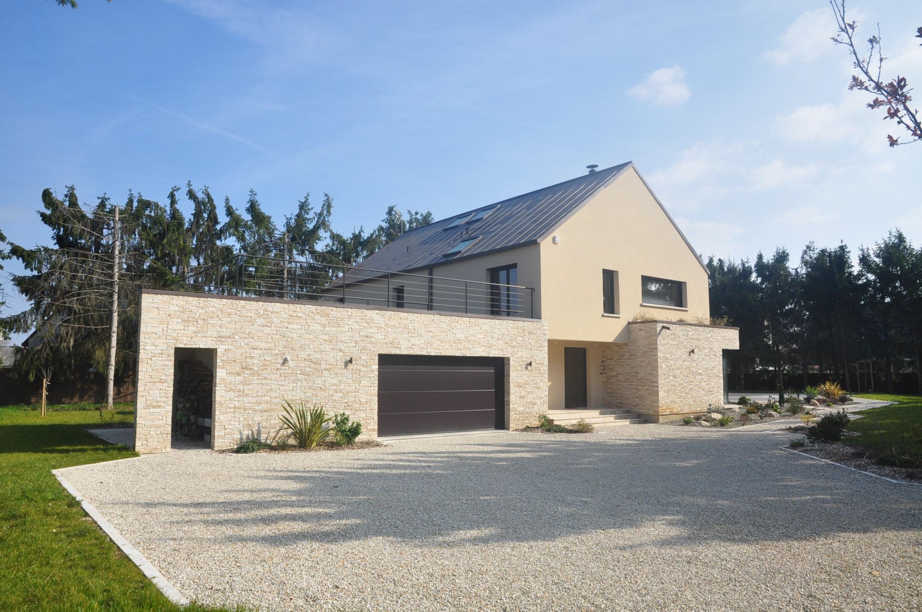 Maison moderne beige et pierre, toit gris, garage et entrée. Allée en gravier clair. Jardin et grands arbres en arrière-plan sous ciel bleu.